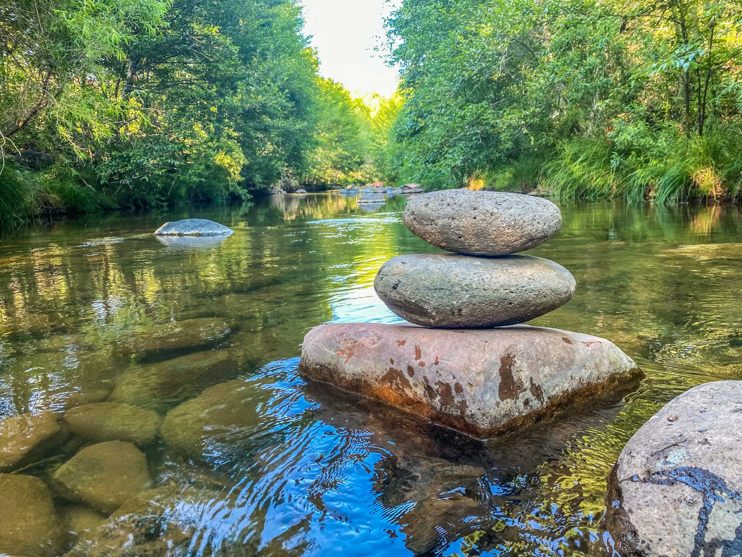 A cairn of three balanced stones rests in a clear, shallow creek, surrounded by the lush green trees of a forest.
