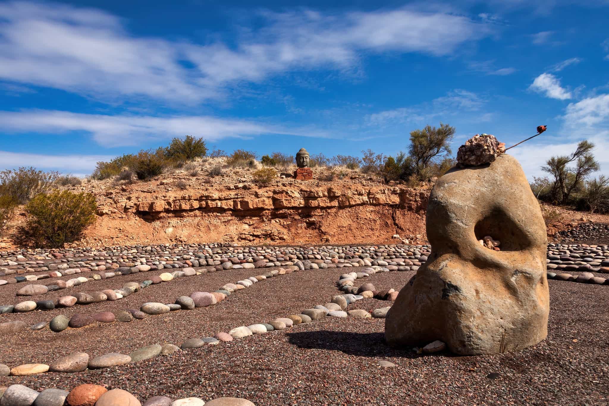 A stone labyrinth in the desert, with a large, uniquely shaped rock in the foreground and a Buddha head statue resting on a red rock ledge in the background under a blue sky.