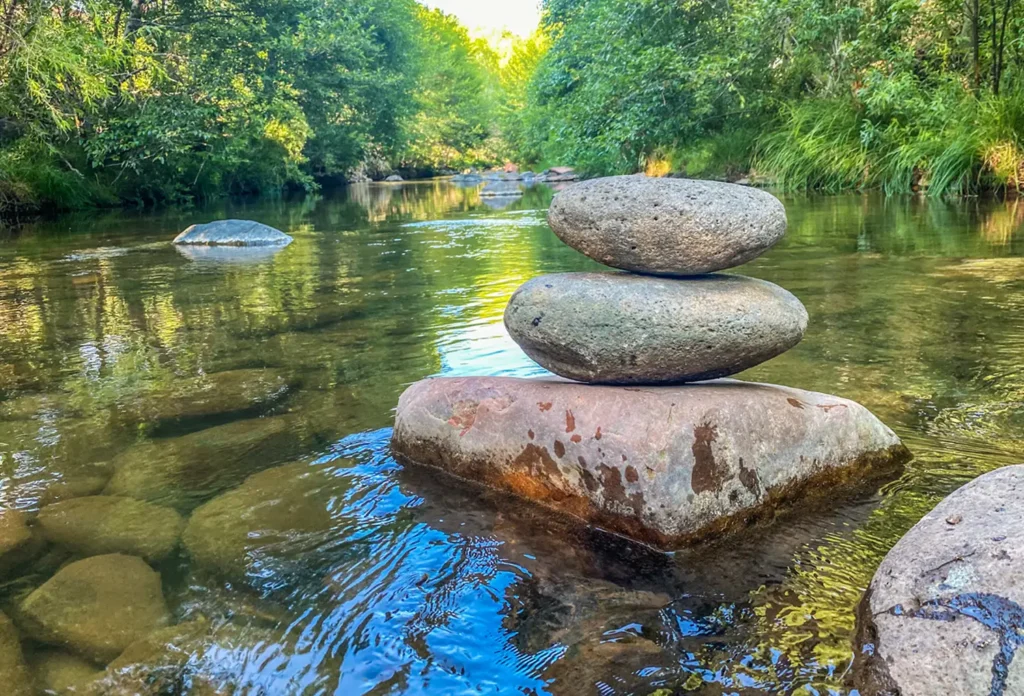 A cairn of three balanced stones rests in a clear, shallow oak creek, surrounded by the lush green trees of a forest.
